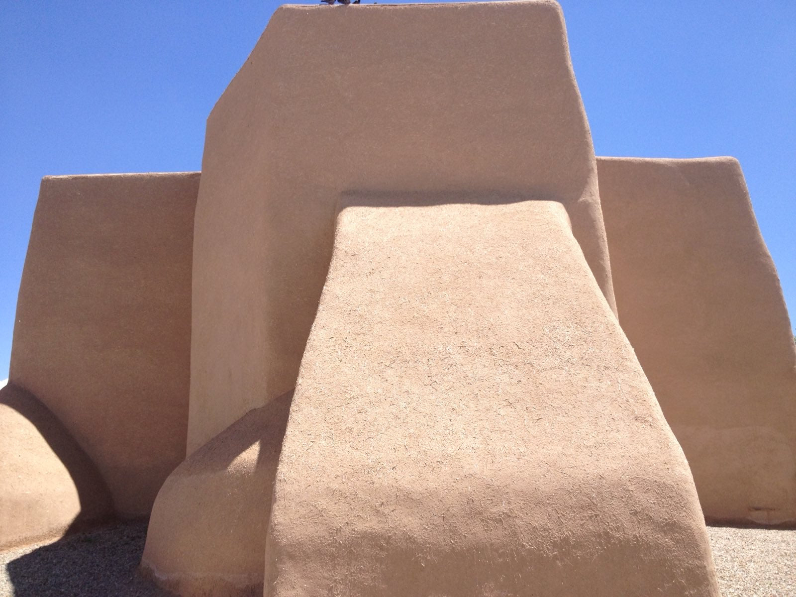 Adobe-style rear view of the San Francisco de Asís Mission Church in Taos, New Mexico, showcasing its traditional smooth, earth-toned architecture under a bright blue sky. The iconic structure reflects the region's rich history and cultural influence, which inspires New Mexico-themed clothing and scarves