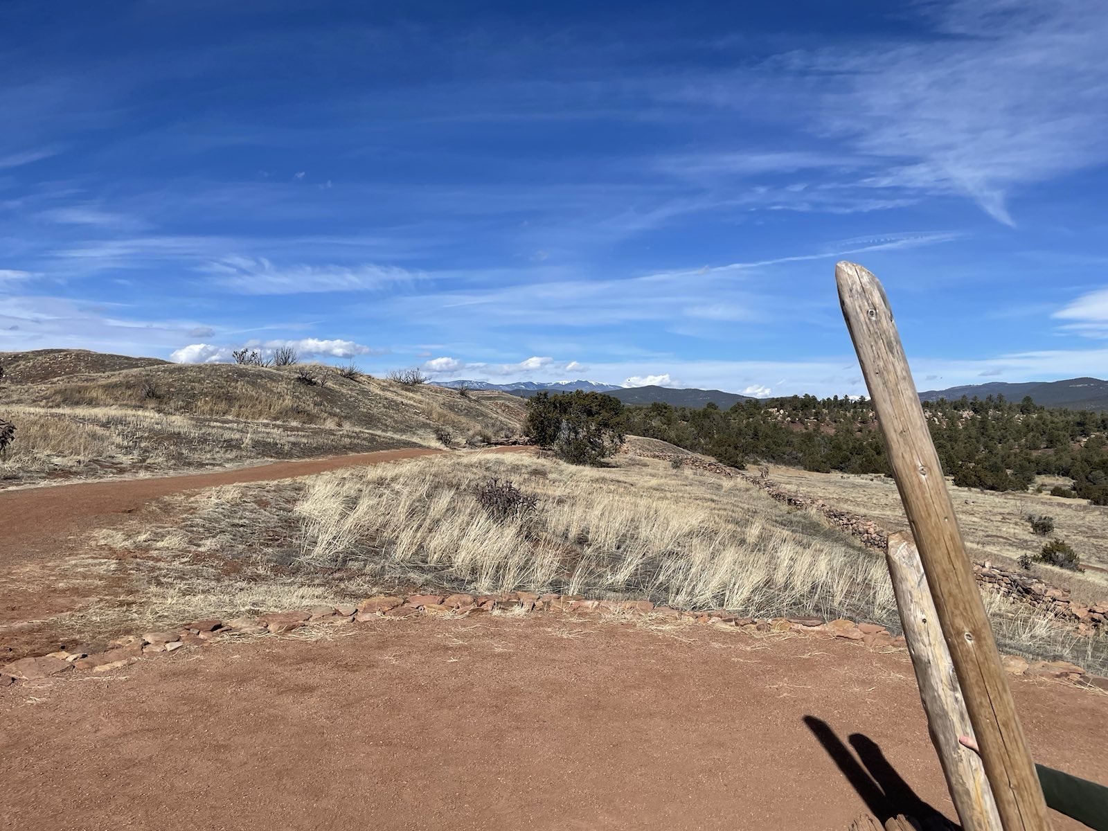 Expansive New Mexico desert landscape with dry grass and red dirt trails winding through rolling hills under a vibrant blue sky. The rugged terrain and natural beauty of the Southwest inspire the clothing designs featured on this site.